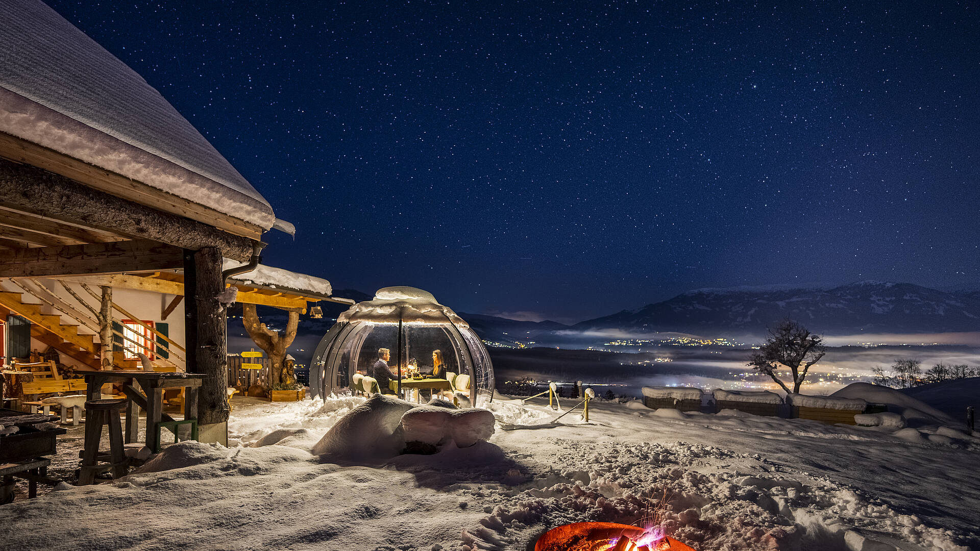 Glaskuppel im Winter von außen, in der ein Pärchen sitzt und das Dinner am Abend genießen
