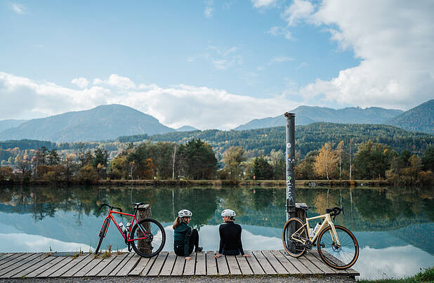 2 Personen rasten am Ufer der Drau mit ihren Gravel Bikes