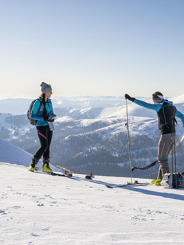 Ein Mann und eine Frau stehen in den Bergen und der Mann zieht gerade die Felle von seinen Tourenskiern ab