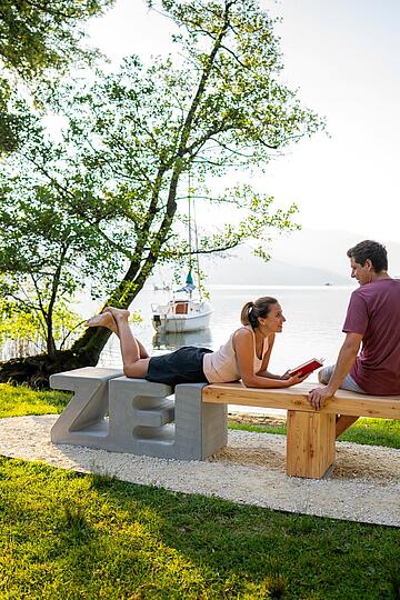Frau liegt auf der Zeitbank und Mann sitzt daneben mit dem Millstätter See im Hintergrund