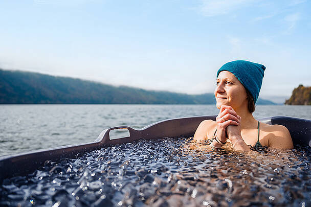 Frau sitzt in einer Holzbadewanne voller Eiswasser und genießt Ausblick auf den Millstätter See