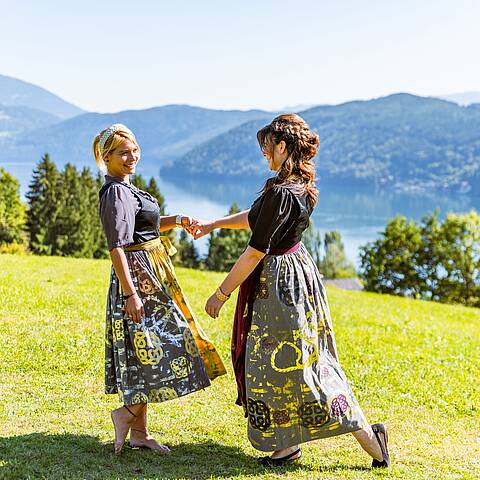 2 Frauen tanzen auf einer Wiese in Tracht mit Blick auf den Millstätter See