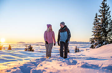 Zwei Personen gehen durch die verschneite Winterlandschaft der Nockberge bei tief stehender Sonne.