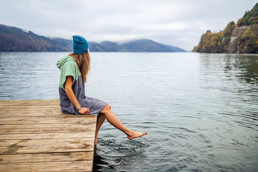 Frau sitzt mit Badeponcho auf Steg am Millstätter See und hält ihre Füße ins Wasser