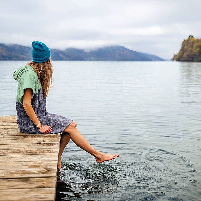 Frau sitzt mit Badeponcho auf Steg am Millstätter See und hält ihre Füße ins Wasser