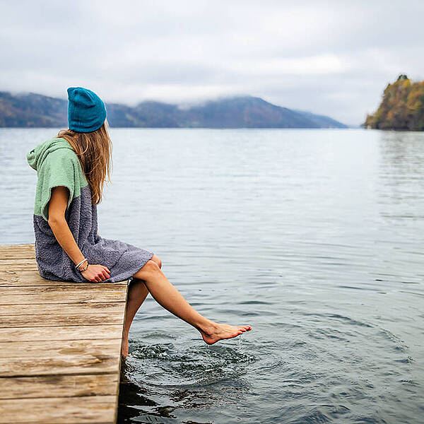 Frau sitzt mit Badeponcho auf Steg am Millstätter See und hält ihre Füße ins Wasser