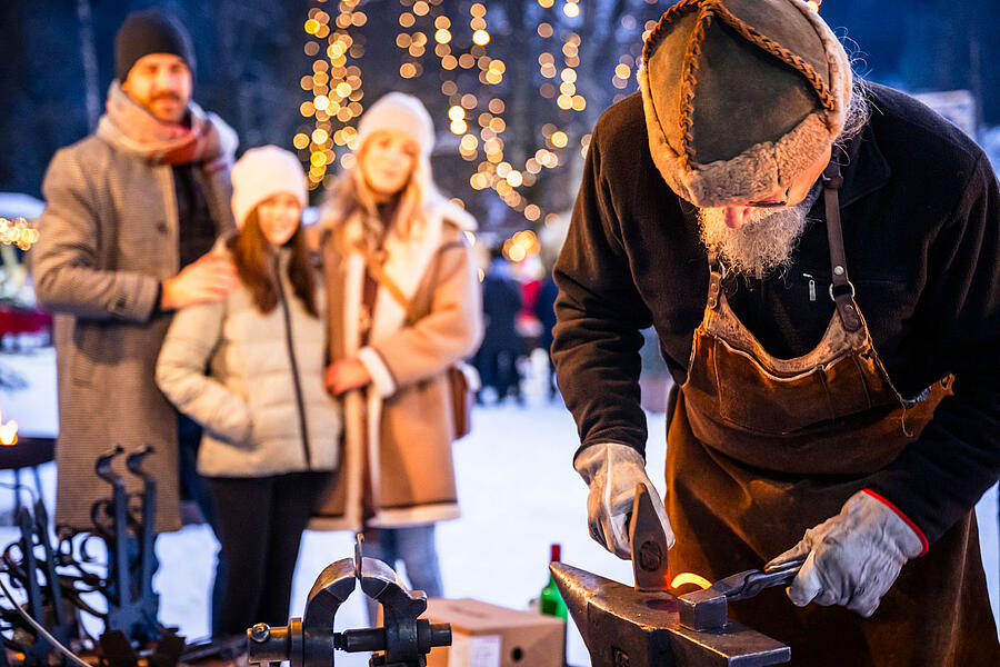 Holzschmied verrichtet seine Arbeit mit einer Familie im Hintergrund am Bad Kleinkirchheimer Advent