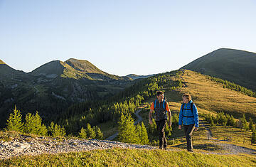  ALT-Text: Familie mit Kindern bei einer Wanderung in den Nockbergen, die glücklich an einem Gipfelkreuz steht und die Aussicht genießt. Bild 2: Wandern in den sanften Nockbergen Bildbeschreibung: Zwei Wanderer, ein Mann und eine Frau, gehen auf einem schmalen Pfad entlang eines grasbewachsenen Bergrückens in den Nockbergen. Der Mann trägt einen orangefarbenen Rucksack und eine graue Weste, die Frau eine blaue Jacke. Sie unterhalten sich und genießen die Bewegung in der Natur. Die Landschaft ist geprägt von sanften, grünen Hügeln und im Hintergrund sind weitere Gipfel der Nockberge zu sehen. Die Sonne scheint und taucht die Szene in ein warmes, goldenes Licht, das die friedliche und idyllische Atmosphäre des Wanderns unterstreicht. ALT-Text: Zwei Wanderer auf einem Wanderweg in den sanften, grasbewachsenen Nockbergen bei sonnigem Wetter.