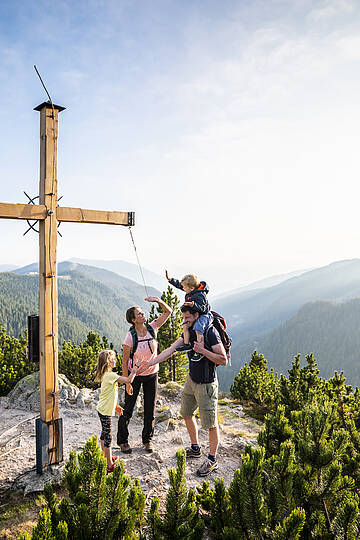 Familie mit Kindern bei einer Wanderung in den Nockbergen, die glücklich an einem Gipfelkreuz steht und die Aussicht genießt.