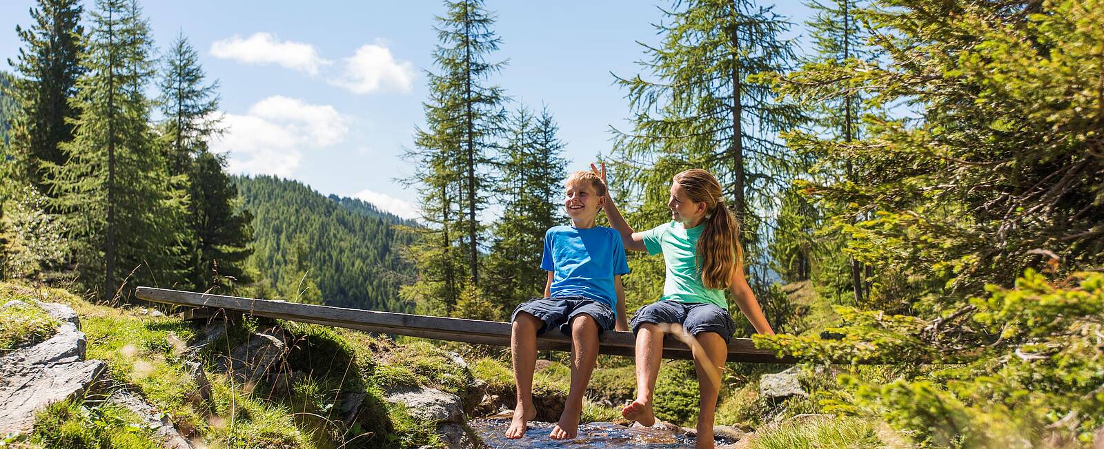 Zwei Kinder sitzen auf einer kleinen Brücke am Bach und genießen die Aussicht auf die Nockberge während einer Wanderung in Bad Kleinkirchheim